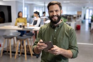 homem sorridente usando tablet em pé no meio de um coworking com pessoas trabalhando em um fundo desfocado
