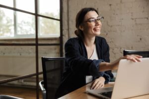 Empreendedora com a mão no topo do notebook, em seu escritório, sorrindo e olhando para além da câmera.