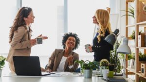 Três mulheres sorrindo e conversando no escritório enquanto tomam café, algumas plantas espalhadas pela mesa e prateleiras.