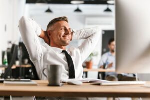 Homem sorridente com as mãos atrás da cabeça relaxa no coworking por estar livre de doenças emocionais.