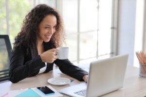 Mulher em escritório home office sorrindo enquanto usa o notebook e toma café.