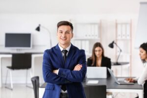 redução de custo​: homem sorridente de braços cruzados com colegas de trabalho ao fundo no escritório